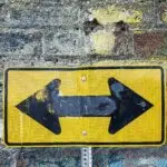 02 Curiosity & Mindset Weathered yellow two-way street sign on a textured rustic brick wall. Urban art style.